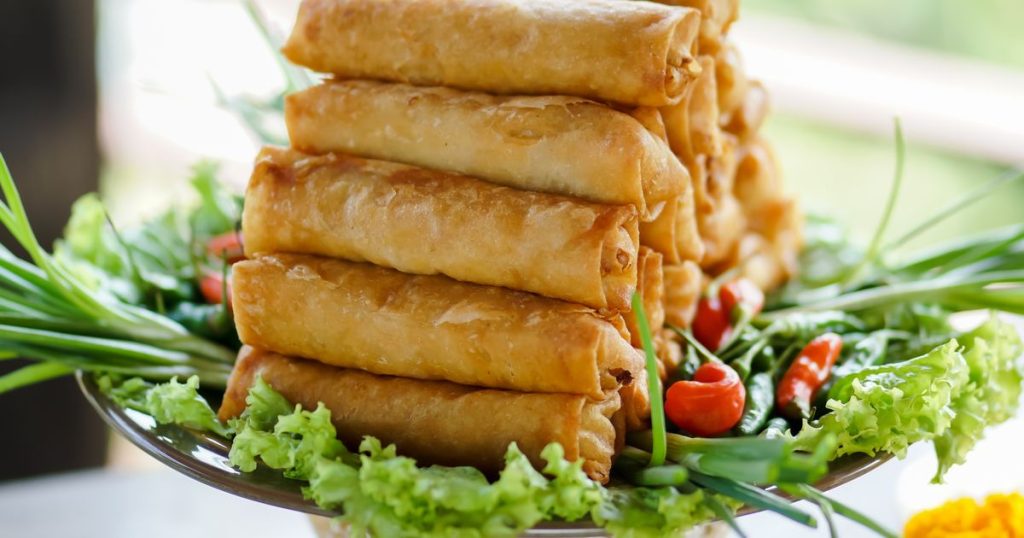Stack of golden crispy spring rolls on a decorative platter with fresh greens and cherry tomatoes