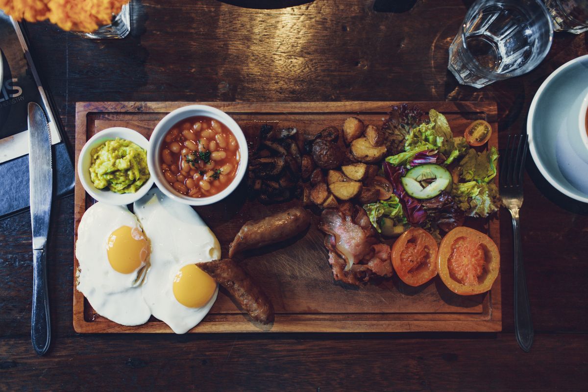 Overhead view of a full breakfast plate with sausage, fried egg, beans, and vegetables on a rustic wooden board